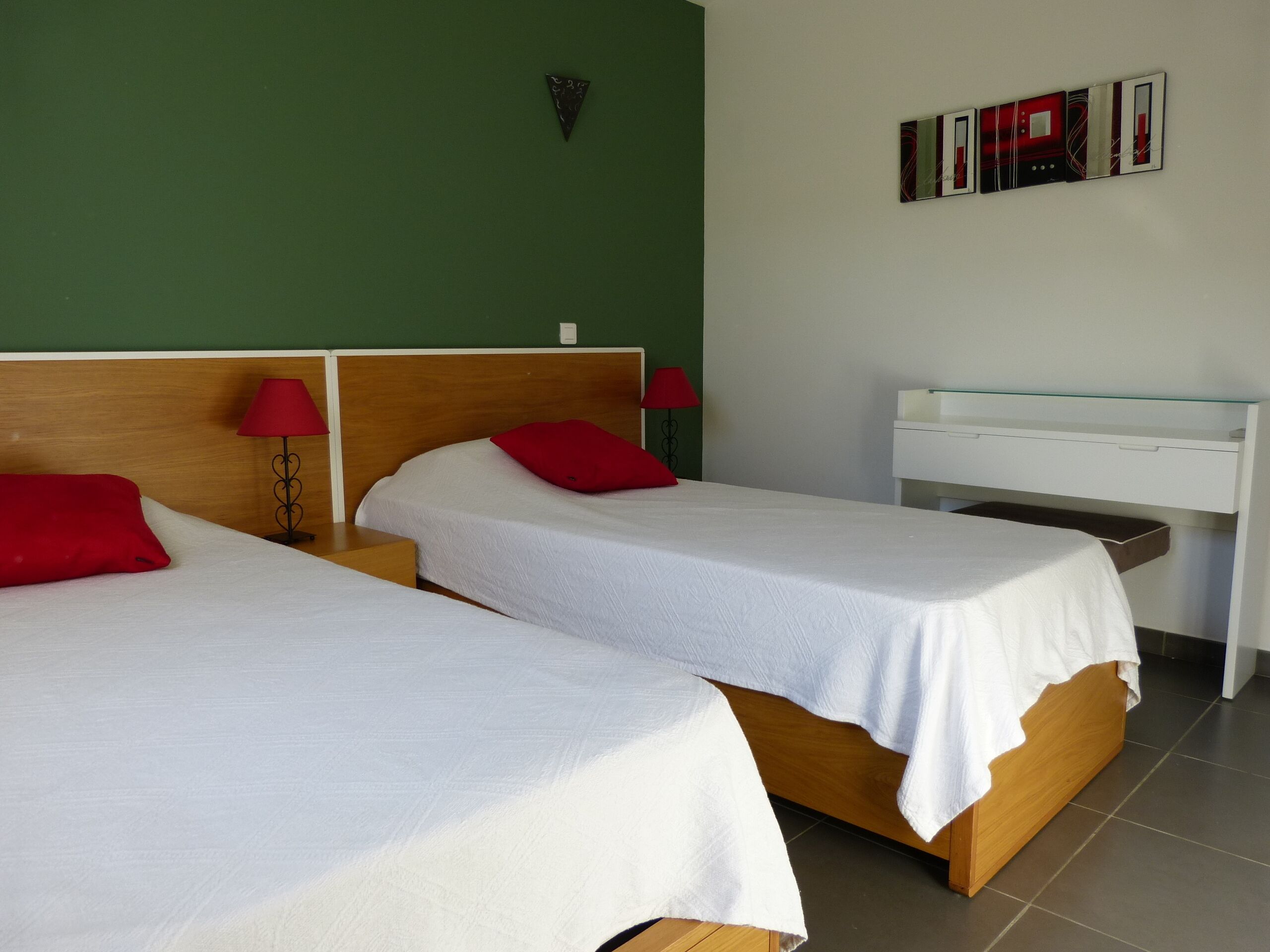 Une chambre d’hôtel avec deux lits simples, chacun agrémenté d’un oreiller rouge et d’un drap blanc. Une petite table de chevet avec une lampe rouge est placée de chaque côté de chaque lit. Un bureau blanc avec un plateau en verre est adossé au mur.