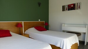 Une chambre d’hôtel avec deux lits simples, chacun agrémenté d’un oreiller rouge et d’un drap blanc. Une petite table de chevet avec une lampe rouge est placée de chaque côté de chaque lit. Un bureau blanc avec un plateau en verre est adossé au mur.