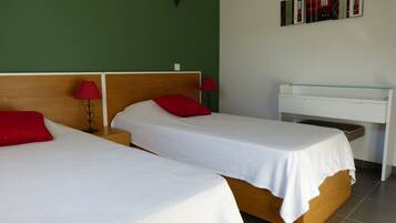 Une chambre d’hôtel avec deux lits simples, chacun agrémenté d’un oreiller rouge et d’un drap blanc. Une petite table de chevet avec une lampe rouge est placée de chaque côté de chaque lit. Un bureau blanc avec un plateau en verre est adossé au mur.