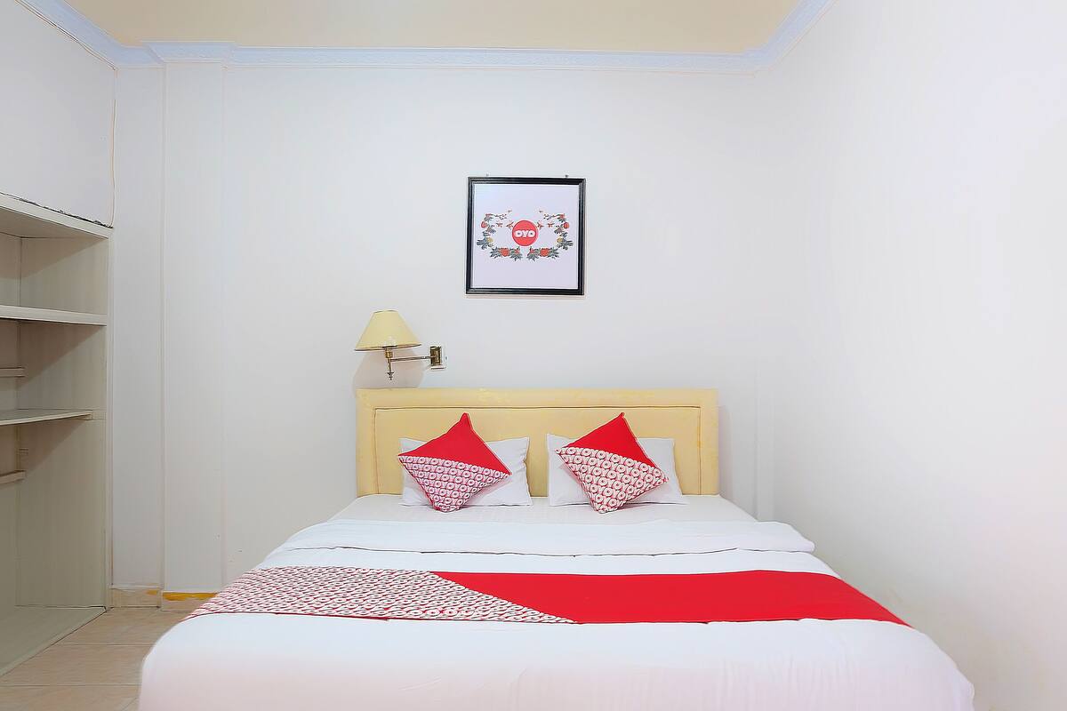 Un lit bien fait, recouvert d’une couvre-lit à motifs rouges et blancs, deux oreillers aux motifs rouges et blancs, un tableau encadré accroché au mur et une lampe de chevet.