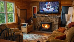 A cozy cabin interior with a stone fireplace, wooden furniture, and a television mounted on the wall.