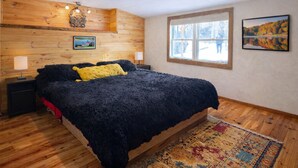 A bedroom with a wooden headboard, a large bed with a dark blanket, two bedside tables with lamps, a window with a view of trees, and a framed picture on the wall.