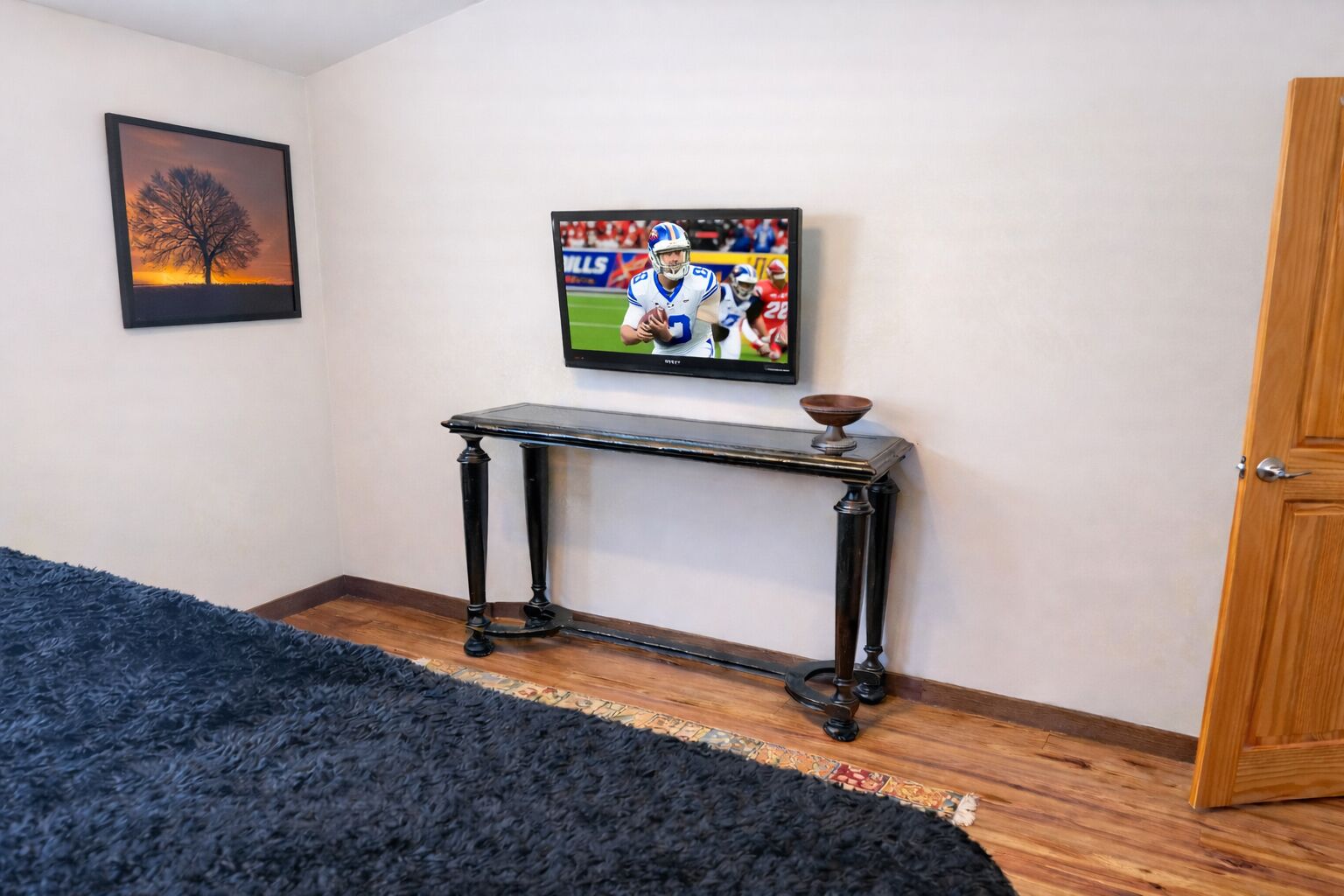 A room with a wooden floor, a black rug, a black console table, a framed picture on the wall, and a door.
