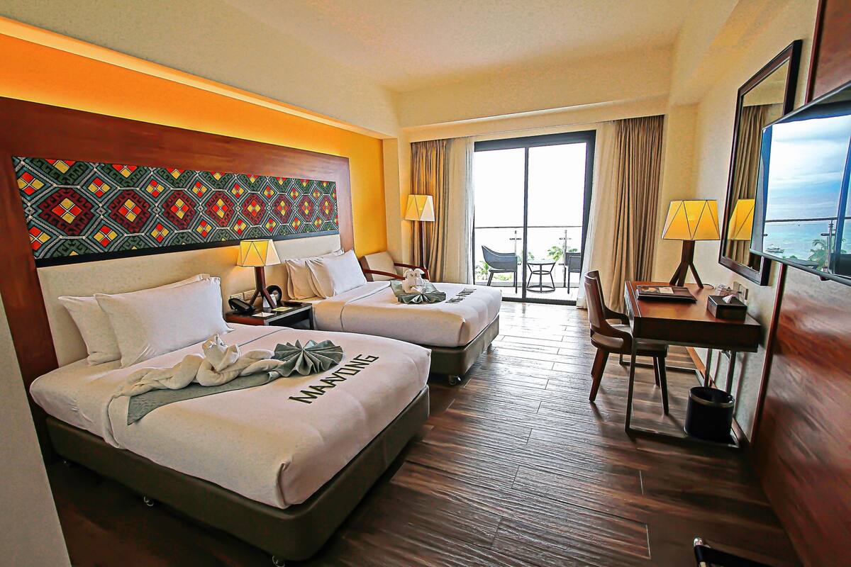 A hotel room with two beds, a desk, a chair, and a balcony with a view.