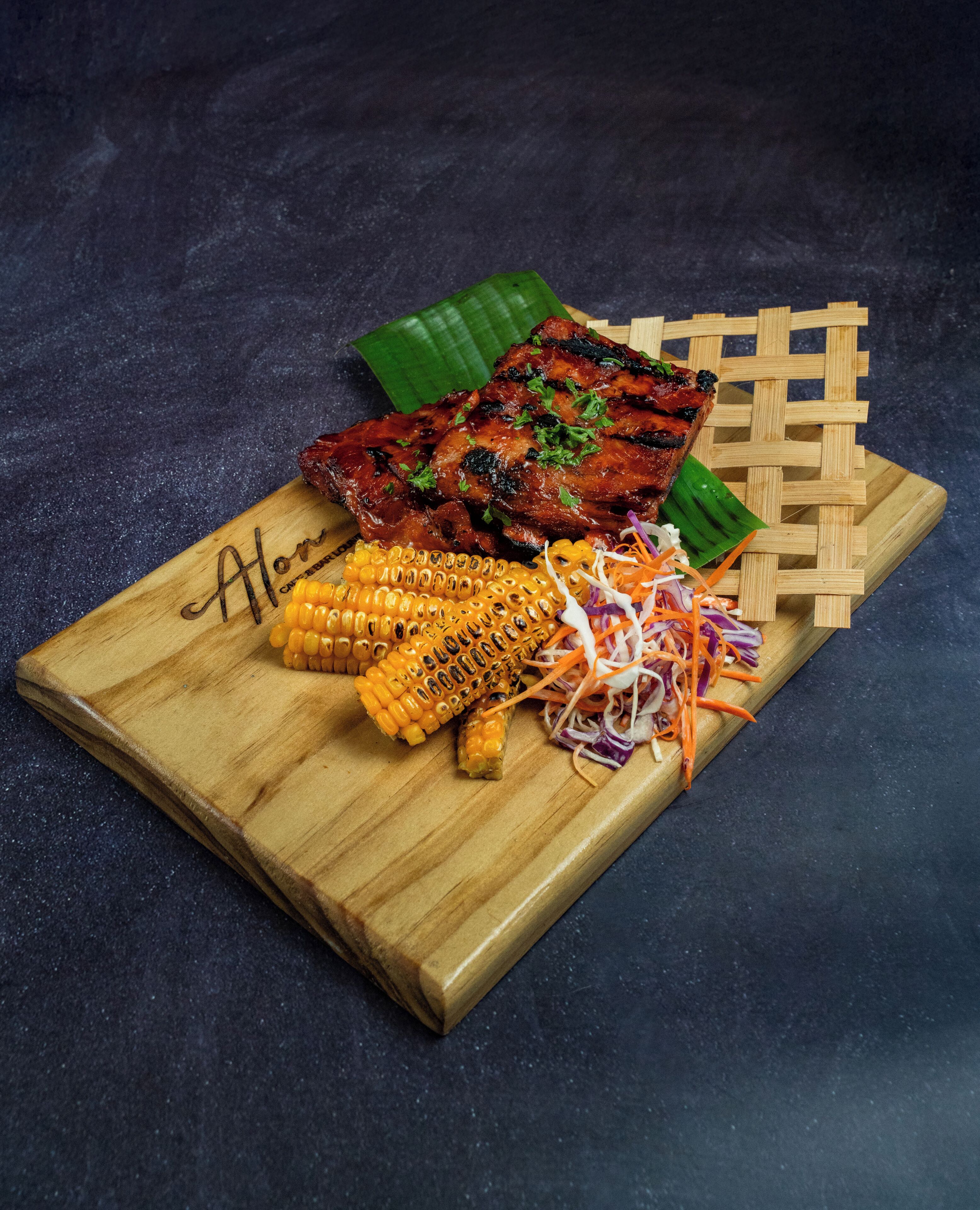 Une planche à découper en bois sur laquelle on voit de la viande grillée, des épis de maïs et une salade de chou, le tout posé sur une surface sombre.