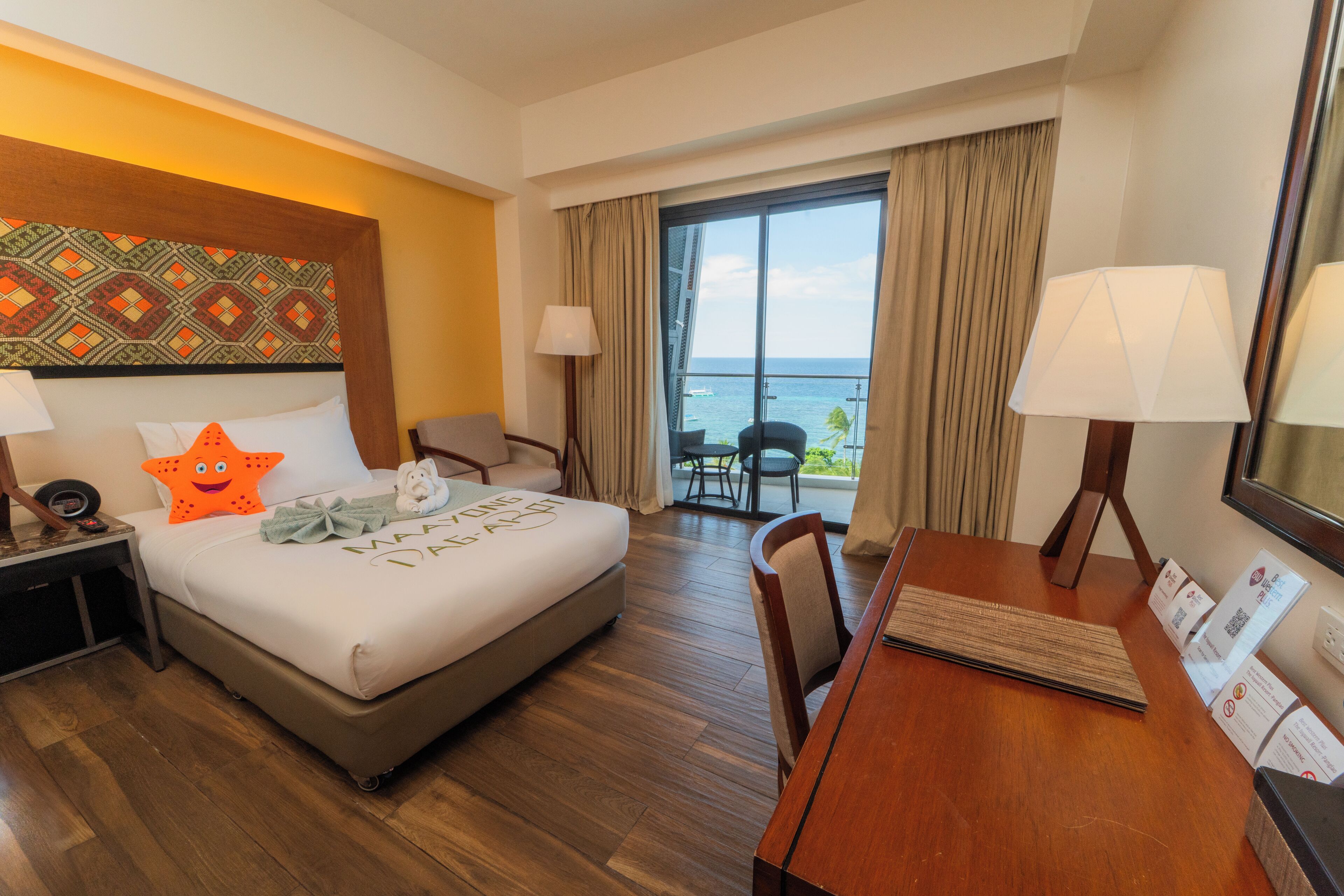 Habitación de hotel con cama, escritorio, silla, lámpara, mesita de noche y vistas al océano a través de amplias ventanas.