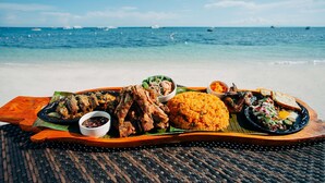 A wooden tray with grilled chicken, rice, and vegetables on a beach with the ocean in the background.
