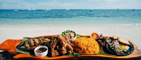 A wooden tray with grilled chicken, rice, and vegetables on a beach with the ocean in the background.