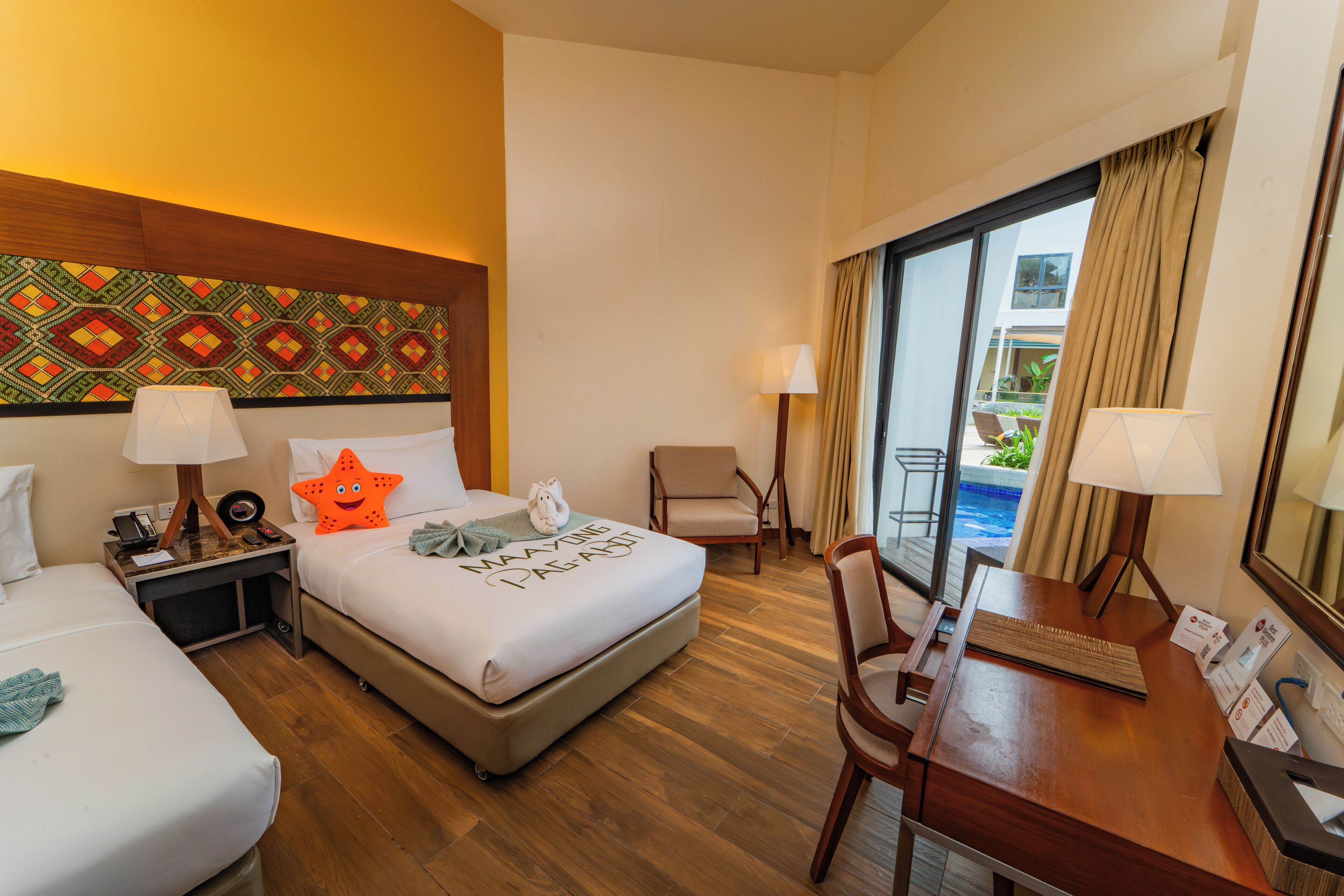 A hotel room with two beds, a desk, a chair, a window with a view of a pool, and a patterned wall.
