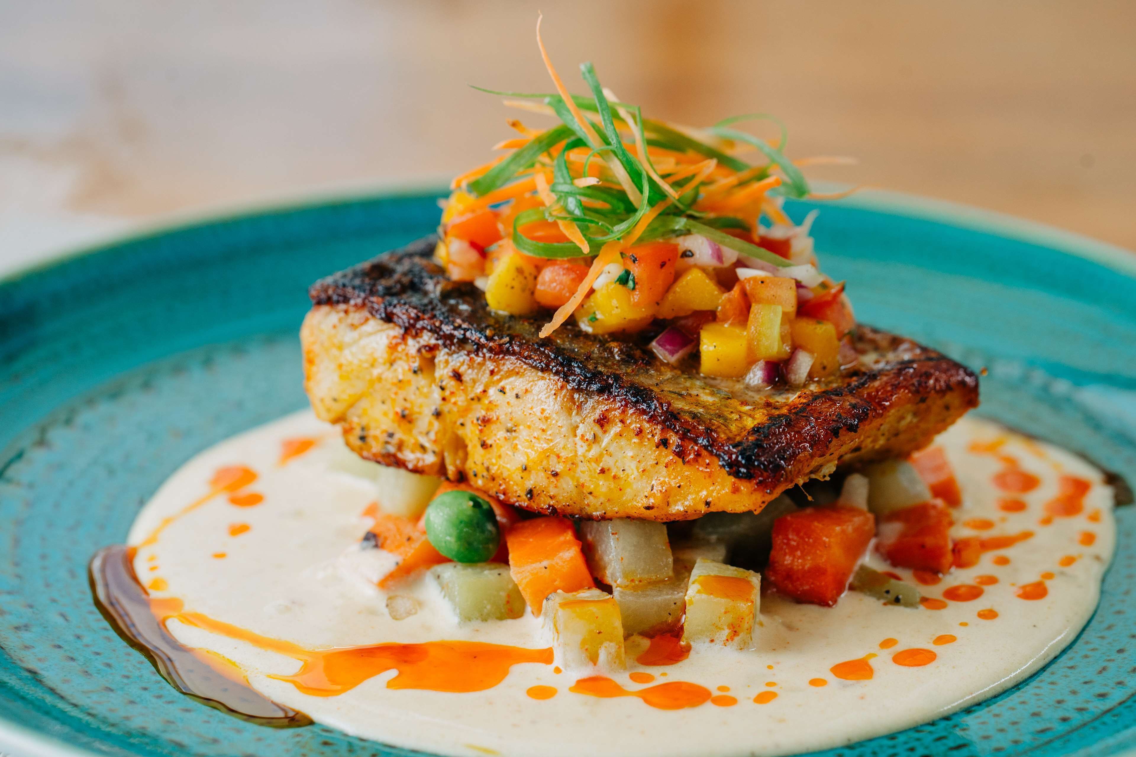 A plate with a grilled fish topped with diced mango and vegetables, served on a blue plate with a creamy sauce.