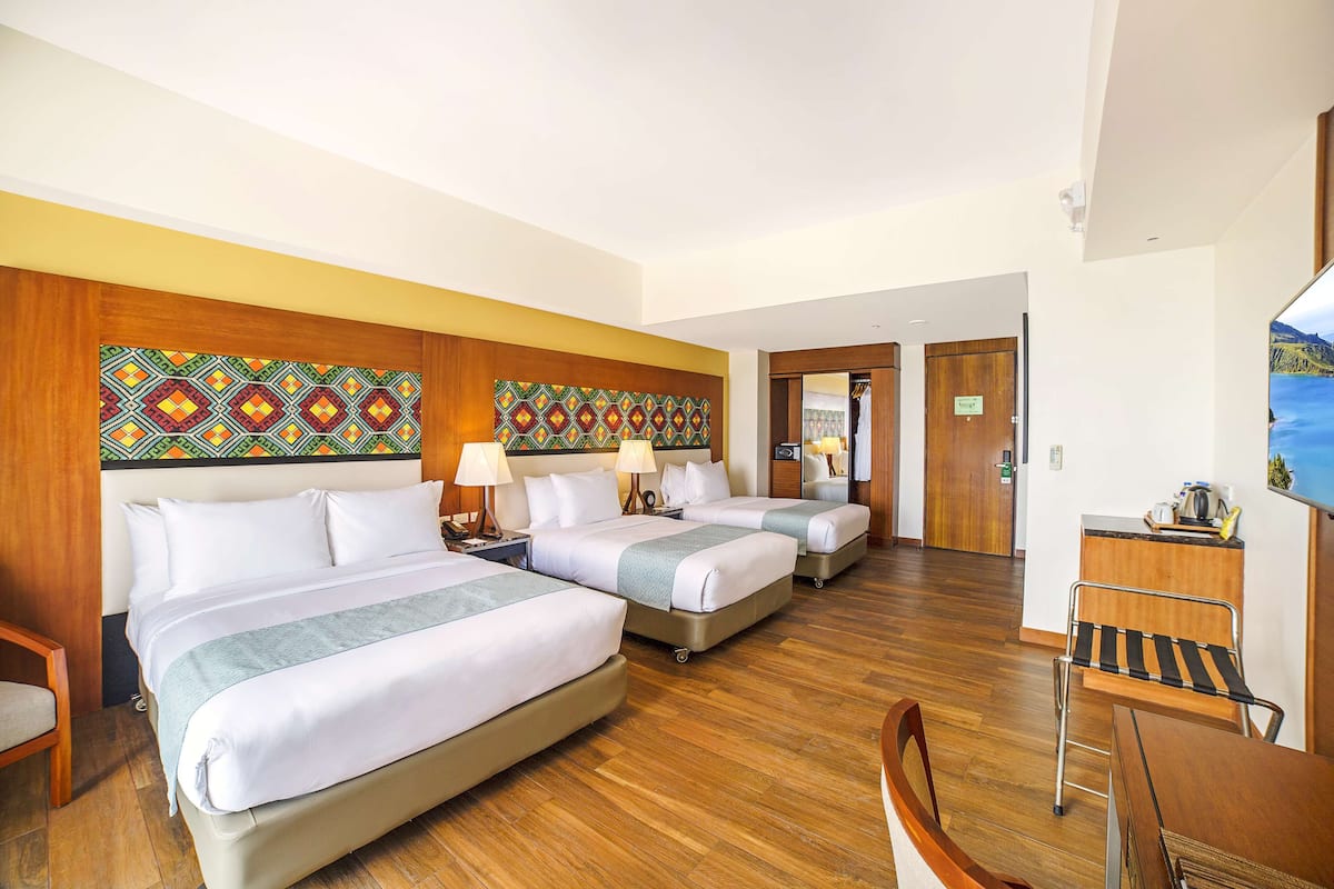A hotel room with three beds, wooden flooring, a desk with a chair, and a television.