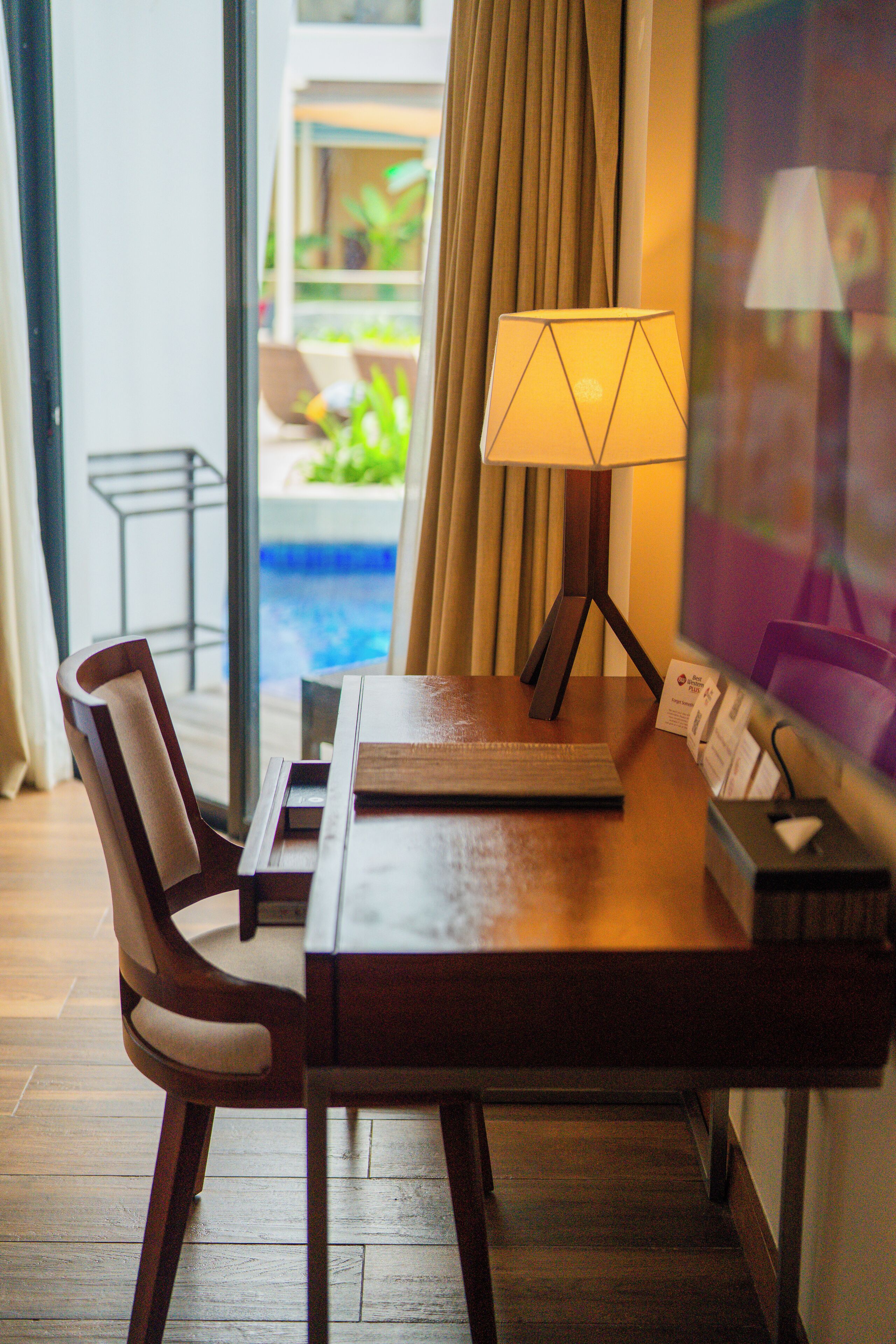 A hotel room with a wooden desk, a lamp, and a view of an outdoor pool.