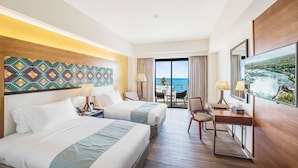 A hotel room with two beds, a desk, a chair, a TV, and a view of the ocean.