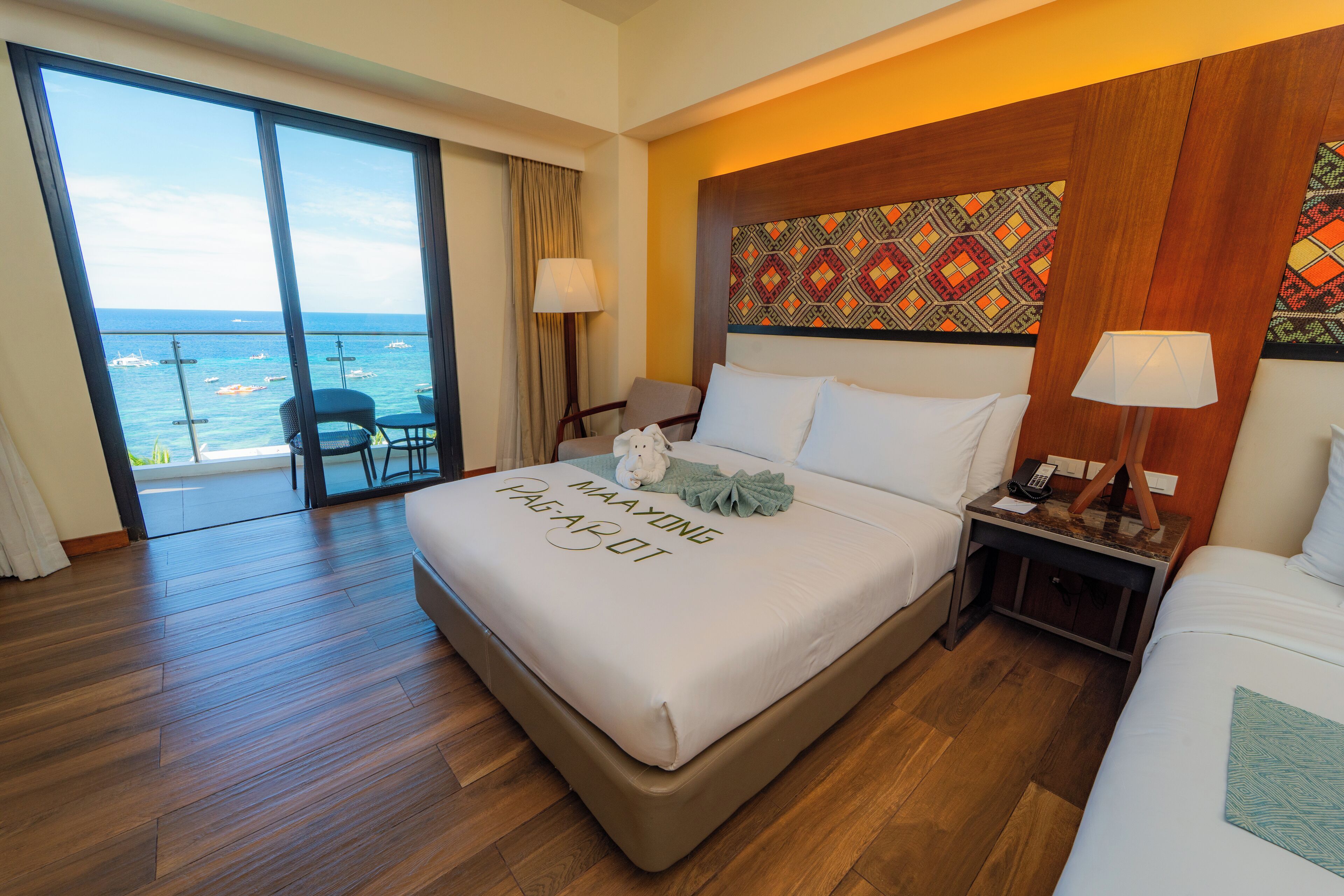 A hotel room with a large bed, a nightstand, a chair, a view of the ocean, and a balcony.