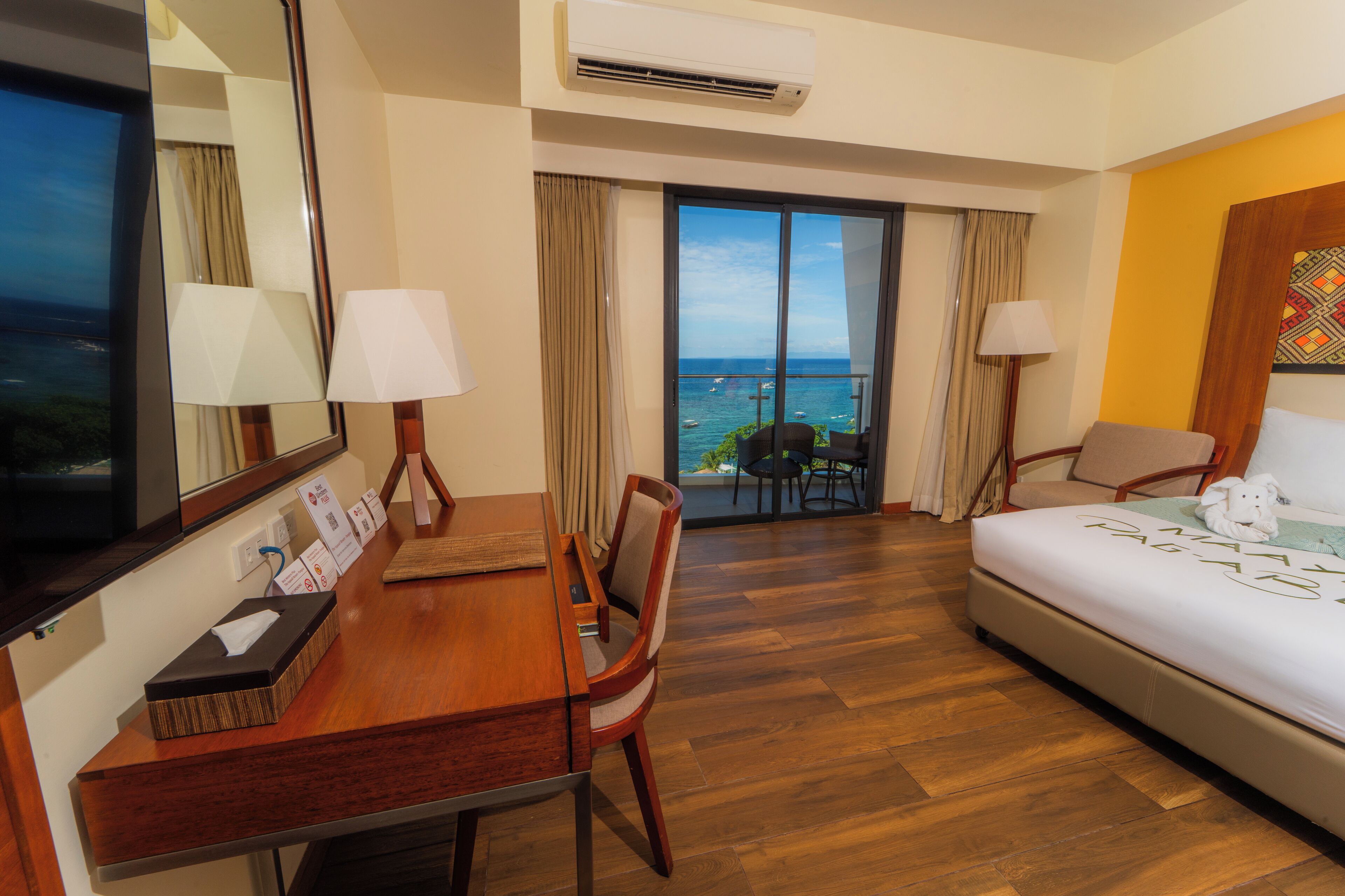 Camera d'albergo con una scrivania in legno, due sedie, un letto e un'ampia finestra vista mare.