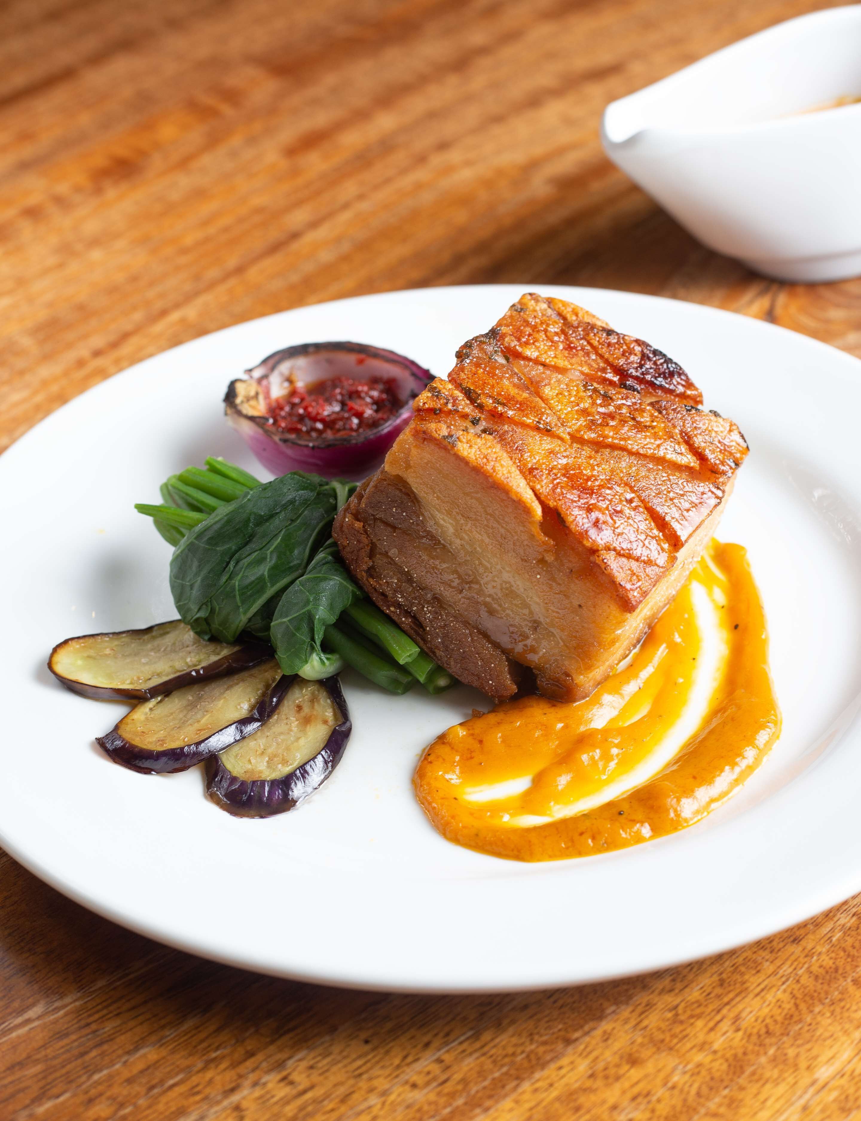 A plate with a glazed pork belly, spinach, eggplant slices, and a sauce.