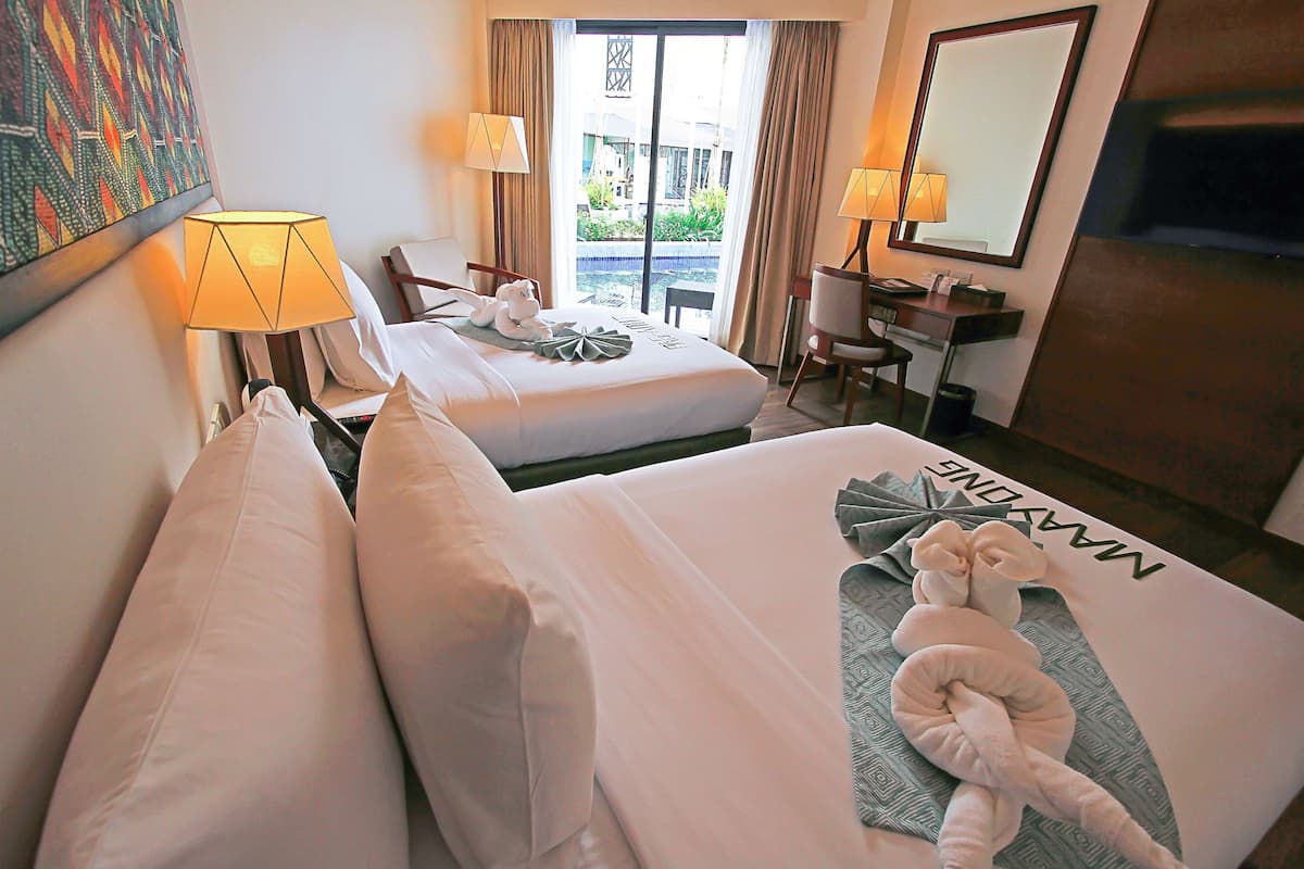 A hotel room with two beds, a desk, a chair, and a view of a pool and building through the window.