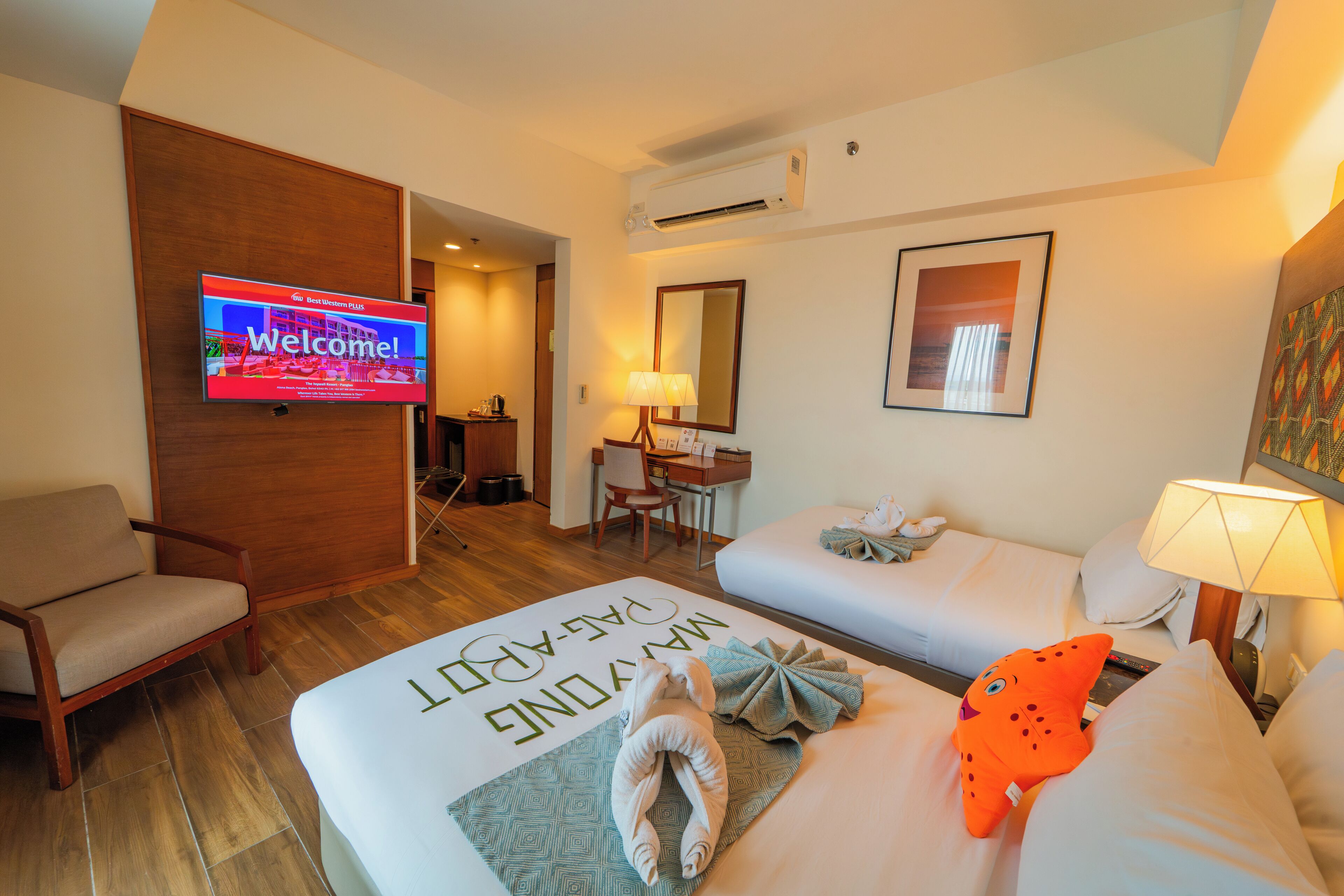 Ein Hotelzimmer mit Bett, Sessel, Schreibtisch, Fernseher, der eine Willkommensnachricht anzeigt, und einer Lampe.