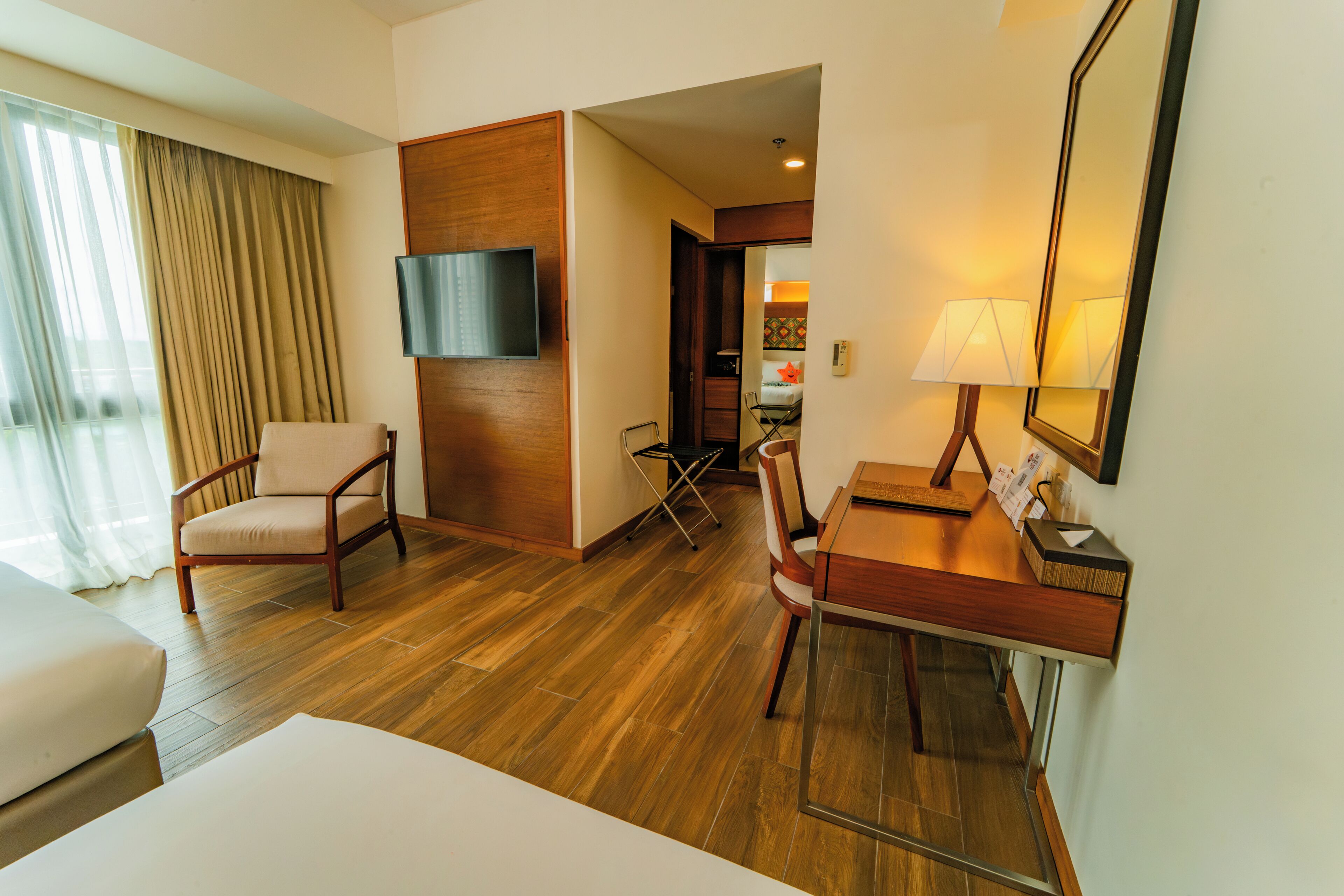 A hotel room with a wooden floor, a desk with a lamp, a chair, a TV mounted on the wall, and a bed.