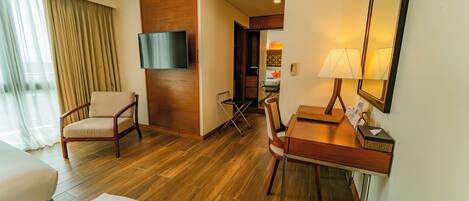 Habitación de hotel con piso de madera, un escritorio con lámpara, una silla, televisor montado en la pared y una cama.