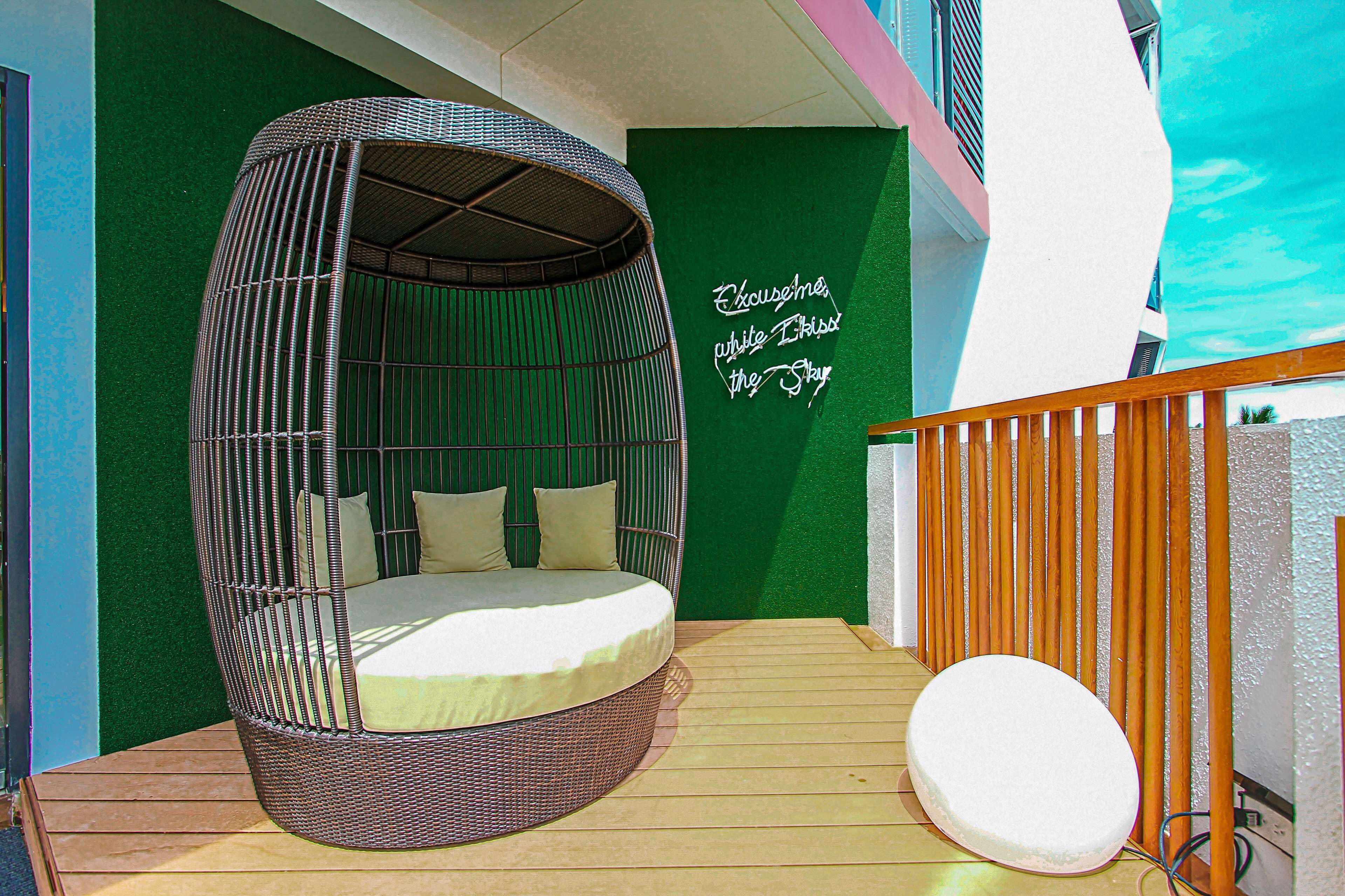 A wicker hanging chair with cushions on a wooden deck, a white cushion on the floor, and a green wall with a sign.