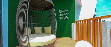 A wicker hanging chair with cushions on a wooden deck, a white cushion on the floor, and a green wall with a sign.