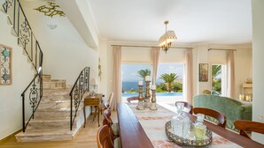 Un vaste espace de vie agrémenté d’un escalier en bois, d’une table à manger mise avec de la belle vaisselle et offrant une vue sur la mer grâce à de grandes fenêtres.
