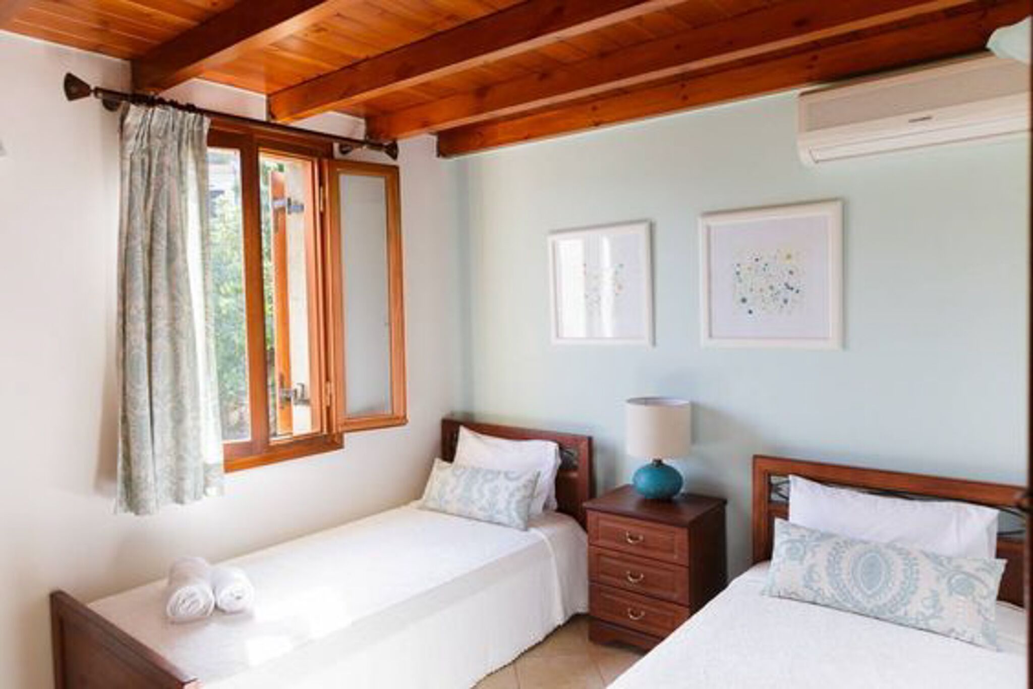 A bedroom with two single beds, a wooden ceiling, a window with curtains, a bedside table with a lamp, and two framed pictures on the wall.