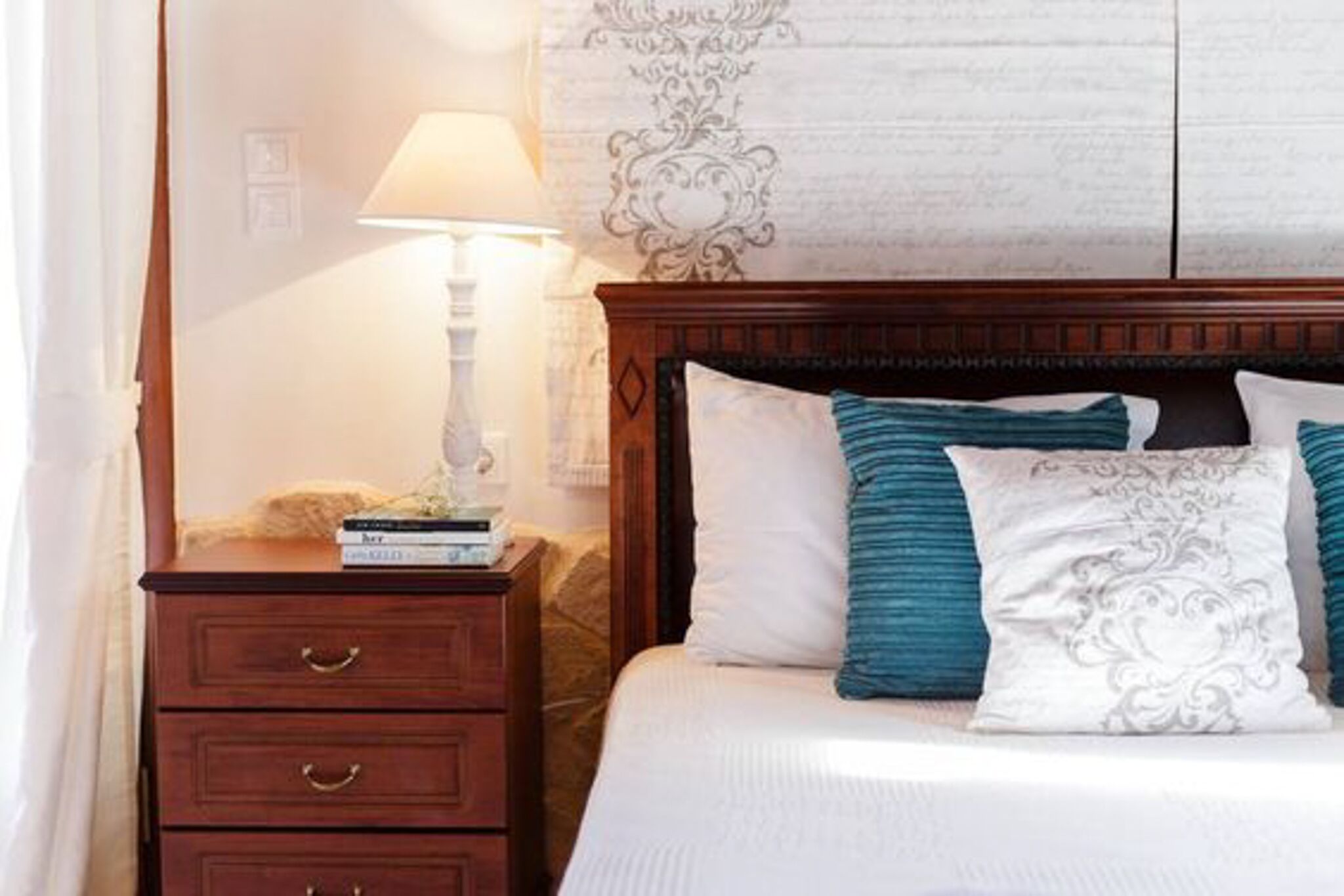 A neatly made bed with white and teal pillows, a wooden nightstand with a lamp, and a patterned wall behind the bed.