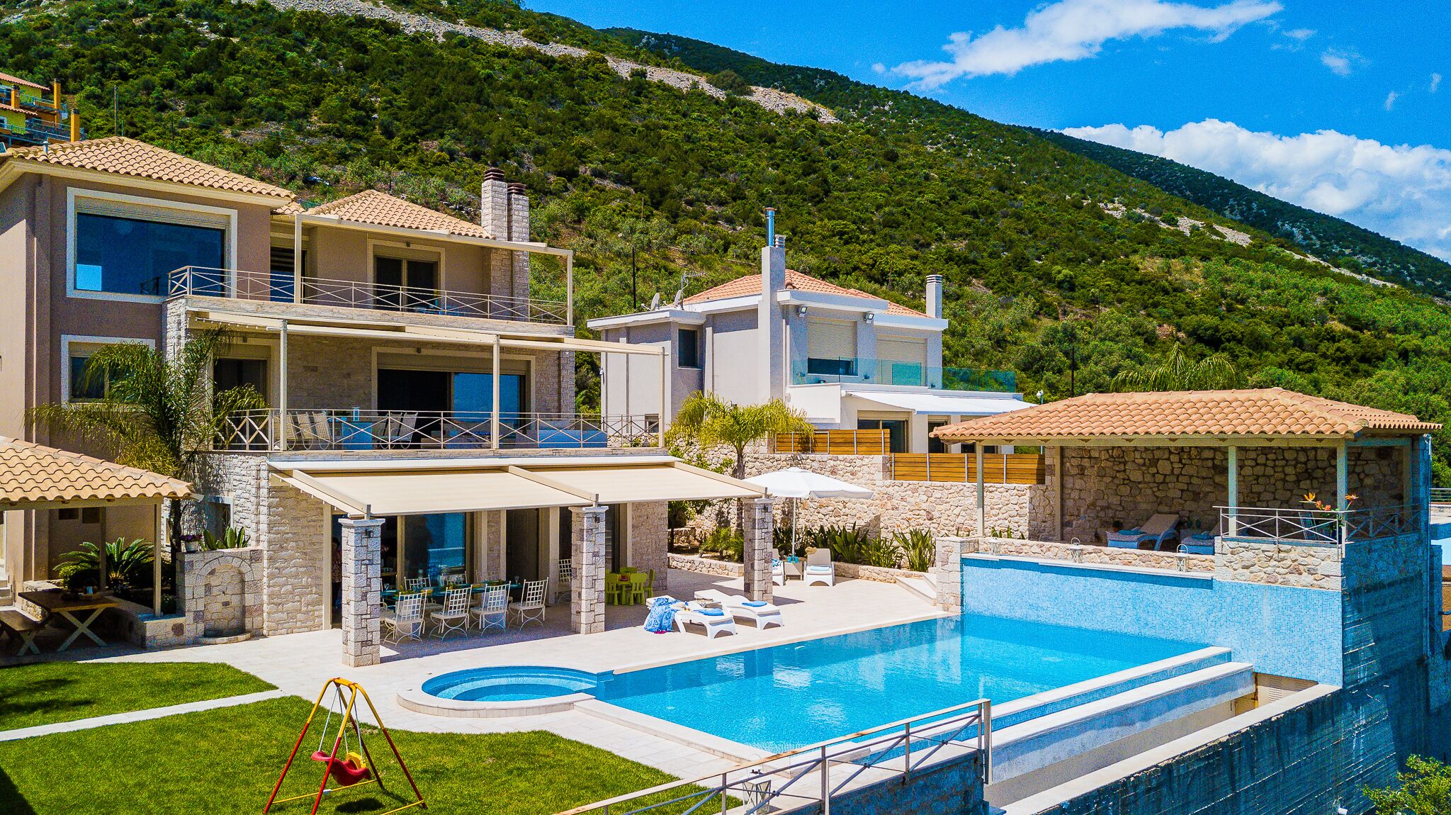 A two-story house with a swimming pool, outdoor seating, and a view of a green hillside.