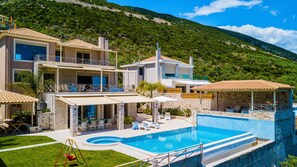 Una casa de dos pisos con piscina, mobiliario exterior y vista a una colina verde.