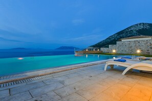Zona de piscina con sillones de descanso y una pared de piedra, con vistas al mar azul turquesa y montañas a lo lejos.