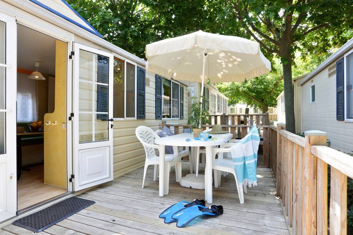 Un espace extérieur abrité, aménagé pour les repas, avec une table et des chaises, un parasol blanc et une paire de chaussures de plage bleues posées sur la terrasse.