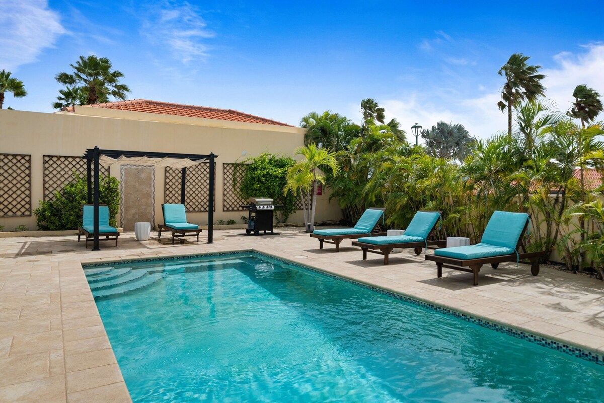 Una piscina con sillones de descanso y una pérgola, rodeada de palmeras y un cielo despejado.