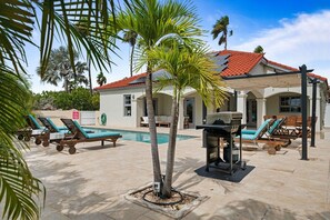 Un área de piscina con sillones de descanso, una parrilla y una palmera.