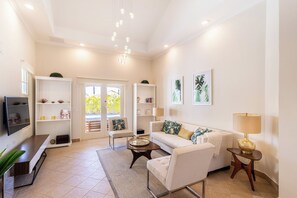 Una sala de estar con un sofá, sillones, una mesa de centro y un televisor. Hay estanterías con objetos decorativos y una puerta que da al exterior.