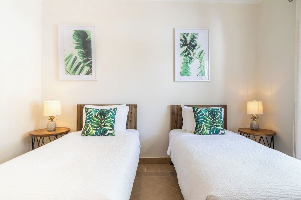 A hotel room with two beds, each with a wooden headboard and a white bedspread. There are two wall-mounted lamps on either side of the beds, and two framed tropical leaf paintings above the beds.