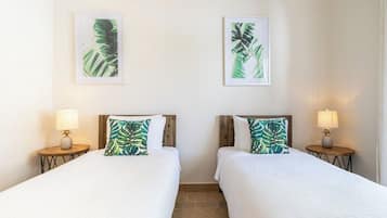 A hotel room with two beds, each with a wooden headboard and a white bedspread. There are two wall-mounted lamps on either side of the beds, and two framed tropical leaf paintings above the beds.