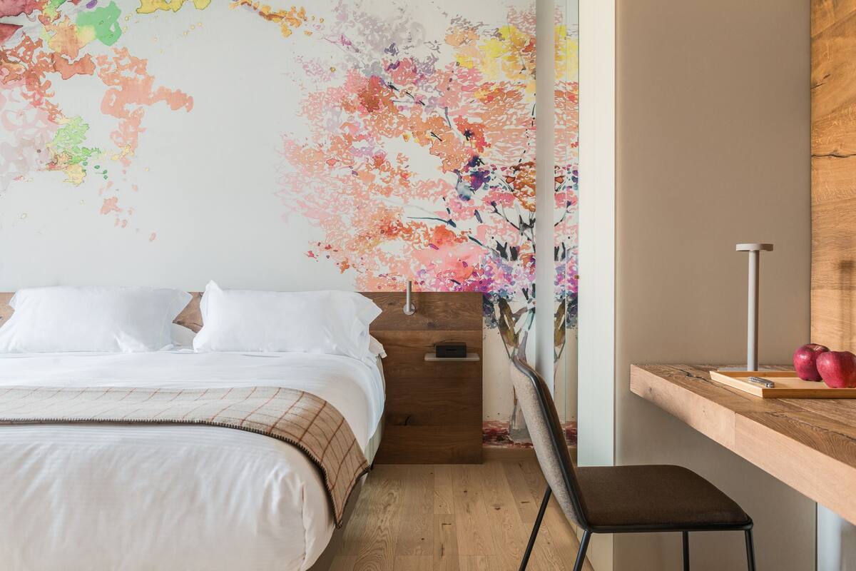 A bedroom with a bed, a wooden nightstand, a desk with a chair, and a colorful floral mural on the wall.