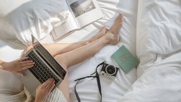 Une personne allongée sur un lit, avec un ordinateur portable, un livre et un appareil photo.