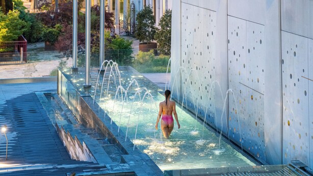Ein modernes Gebäude mit einer reflektierenden Fassade, einem Pool mit Wasserspielen und einer Person im Badeanzug.
