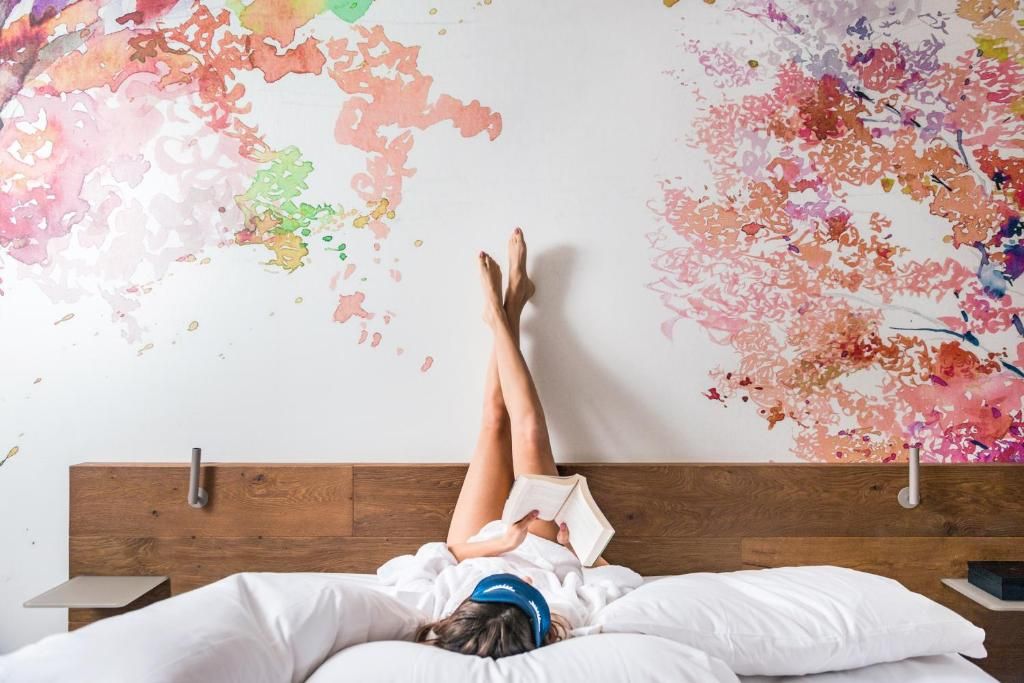 Eine Person liegt auf einem Bett, im Hintergrund ist ein farbenfrohes, abstraktes Wandbild zu sehen.