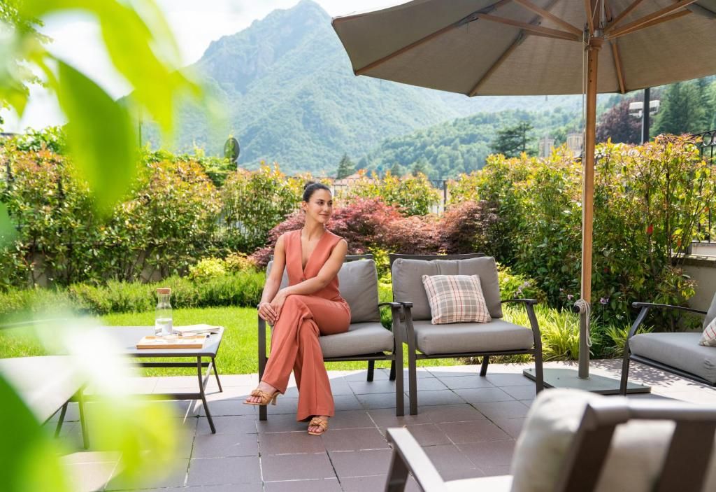 Una donna seduta su un divano grigio con un cuscino, un tavolo con un bicchiere e un libro, e un ombrello in un giardino sullo sfondo di montagne.
