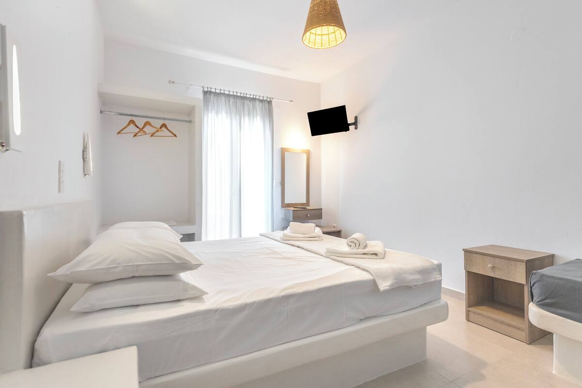 Un dormitorio moderno con una cama grande, una mesita de noche, un televisor montado en la pared y una ventana con cortinas.
