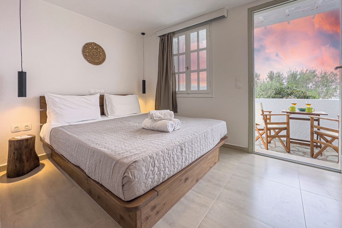 Un dormitorio con una cama grande, una mesita de noche, una lámpara de pared, una ventana con cortinas y vistas al exterior.