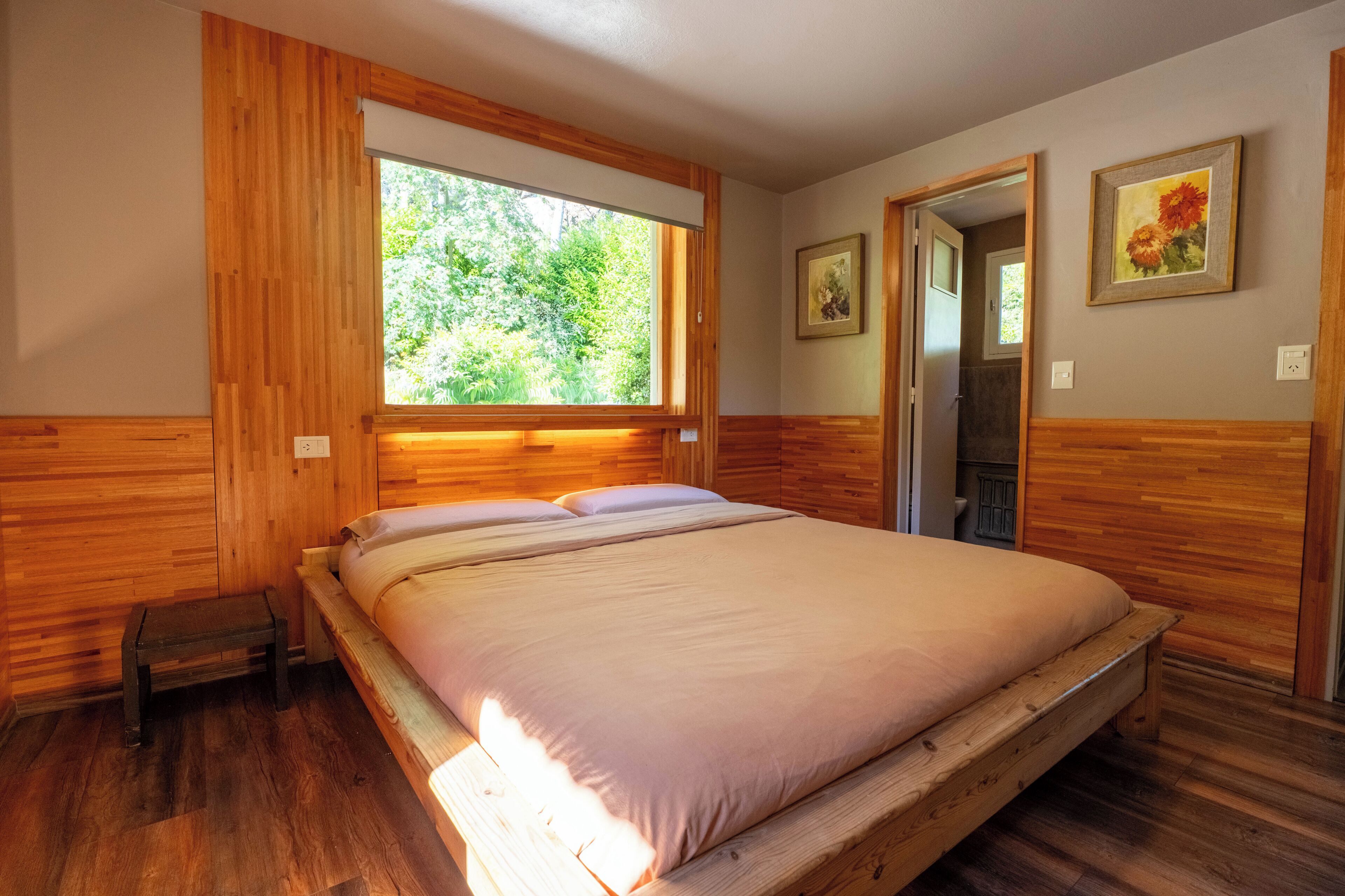 A wooden-paneled bedroom with a large window, a bed, a small bench, and two framed pictures on the wall.