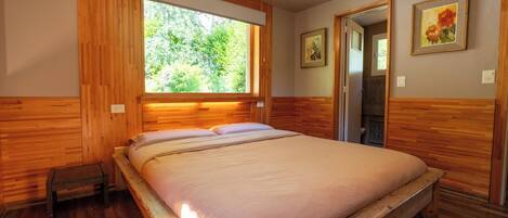 A wooden-paneled bedroom with a large window, a bed, a small bench, and two framed pictures on the wall.