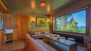 Un intérieur moderne avec un banc en bois, un canapé et une table à manger, une grande fenêtre donnant sur des arbres et l’eau, et un tableau accroché au mur.