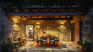 A stone-walled patio with a wooden table and chairs, a hanging light fixture, and potted plants. - Cottage with private pool (Briteiros São Salvador- Guimarães)