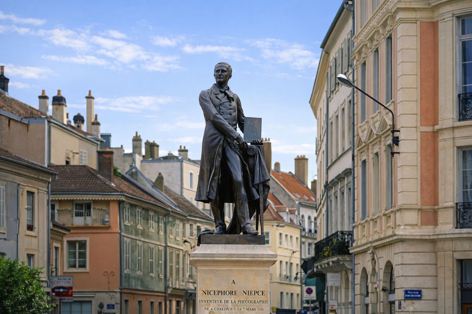 A statue of Nicephore Niepce on a pedestal with a plaque, set against a backdrop of historic buildings.